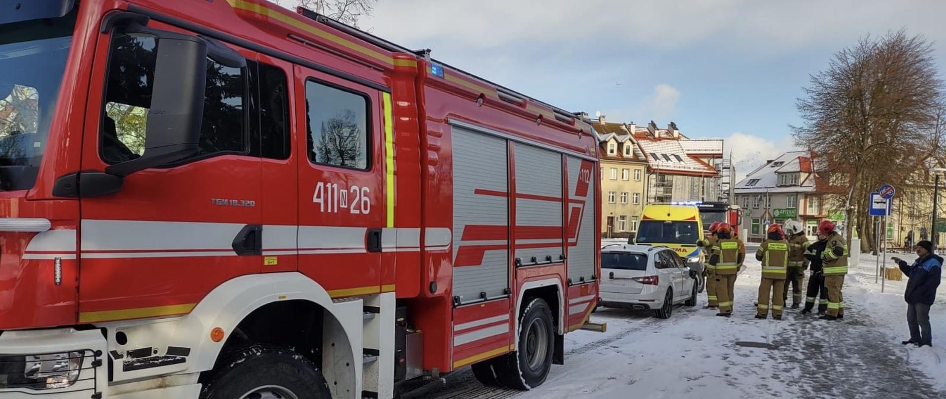 Na zdjęciu na poboczu drogi duży czerwony samochód strażacki, za nim w oddali białe auto osobowe i żółta karetka. Na chodniku z prawej strony strażacy. Biało od śniegu.