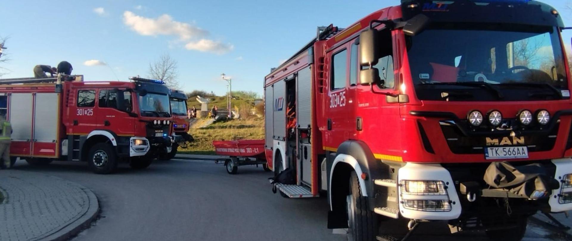 Zdjęcie przedstawia dwa samochody pożarnicze, w tym jeden z podpiętą łódką, zaparkowane przy Rezerwacie Skalnym Ślichowice, który widać w oddaleniu.