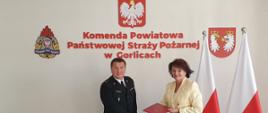 Na zdjęciu Komendant Powiatowy PSP w Gorlicach w umundurowaniu wyjściowym granatowym przekazuje dokument podpisanego porozumienia Pani Dyrektor Powiatowej Stacji Sanitarno-Epidemiologicznej w Gorlicach. Pani Dyrektor ubrana w kremowy żakiet i biała spódnicę. W.w osoby na tle ściany z napisem koloru czerwonego Komenda Powiatowa PSP w Gorlicach n ścianie znajdują się godło Rzeczypospolitej Polskiej, Powiatu i PSP po prawej stronie stoją biało czerwone flagi.