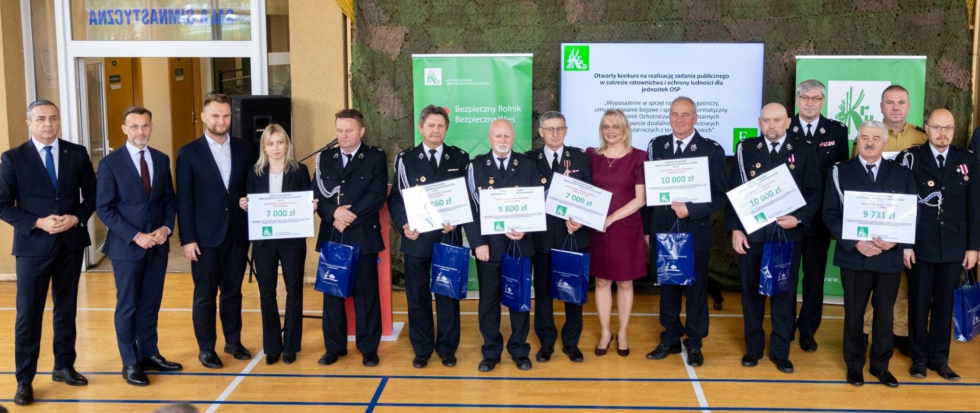 Grupa uczestników stoi na sali gimnastycznej podczas uroczystości przekazania voucherów dla jednostek OSP. W pierwszym rzędzie przedstawiciele instytucji i druhowie w galowych mundurach trzymają otrzymane vouchery oraz torby z upominkami. W tle widoczne banery KRUS i ekran z prezentacją.
