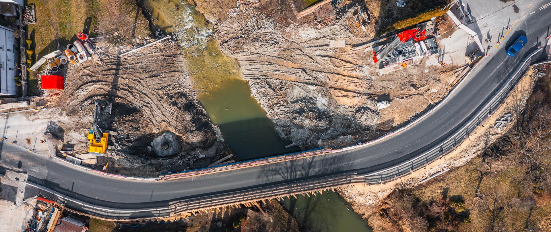 Fotografia przedstawia plac budowy uchwycony z perspektywy lotu ptaka. Centralnym punktem zdjęcia jest rzeka, która przepływa przez środek kadru, dzieląc teren na dwie części. Po lewej stronie rzeki widoczna jest żółta koparka pracująca na ziemi, podczas gdy po prawej stronie dominuje duży, czerwony dźwig oraz inne maszyny budowlane.
Rzeka przecinana jest przez most, który jest zabezpieczony barierkami ochronnymi. W tle widać zabudowania mieszkalne oraz drogi otoczone zielenią, a cały obszar jest otoczony drzewami i roślinnością, co tworzy ciekawy kontrast między naturą a działalnością ludzką.