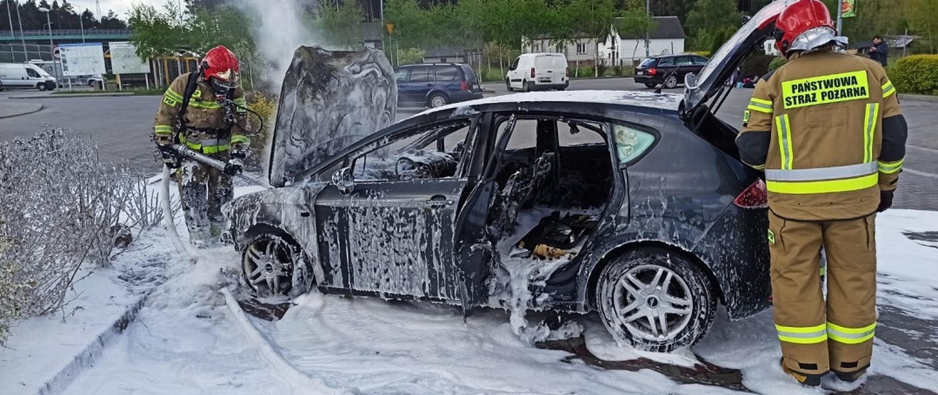 Na zdjęciu widzimy strażaków gaszących palący się samochód osobowy marki Seat. Strażacy ubrani są w ubrania bojowe , strażak ma założony sprzęt ochrony układu oddechowego. Dookoła pojazdu jak i na pojeździe leży piana gaśnicza ciężka.