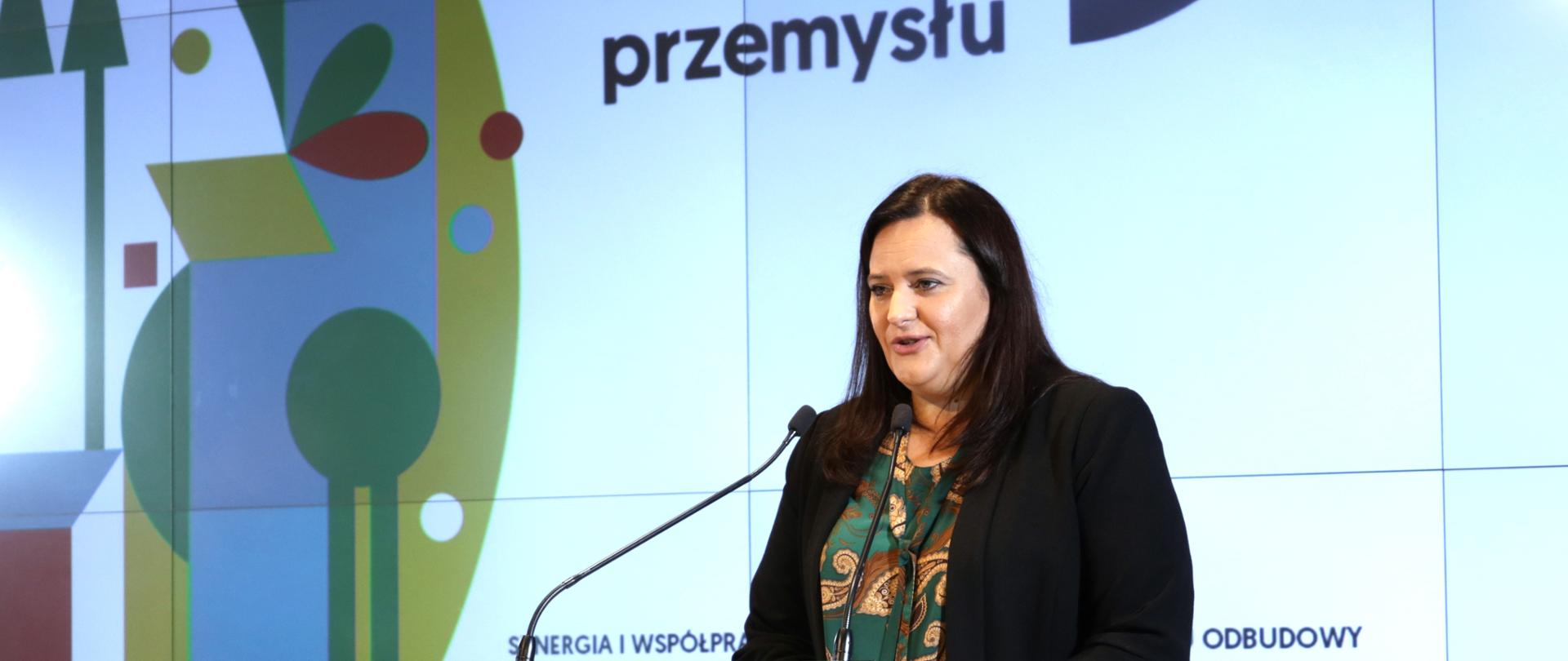 wiceminister Małgorzata Jarosińska-Jedynak mówi do mikrofonu, za nią częściowo widoczny napis Miesiąc Liderów Przemysłu