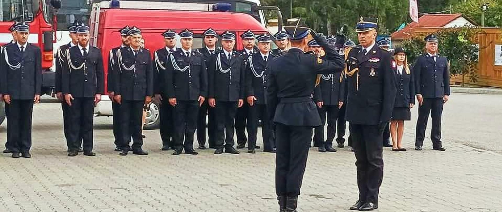 Zdjęcie zrobione na zewnątrz podczas uroczystego apelu. Oficer Państwowej Straży Pożarnej w Stopniu starszego brygadiera odbiera meldunek rozpoczynający uroczystość. W tle ustawieni w dwuszeregu strażacy uczestniczący w apelu.
