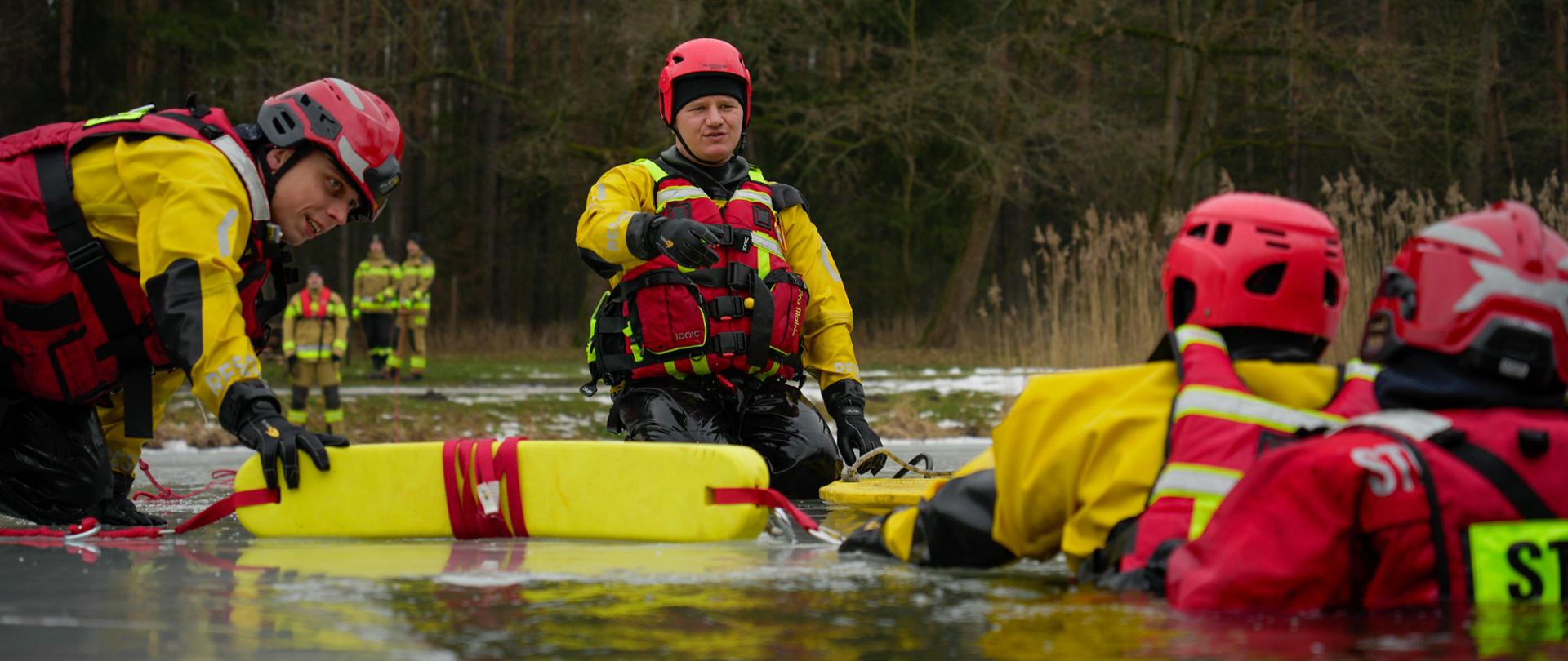 Zdjęcie przedstawia grupę strażaków w specjalnych kombinezonach wypornościowych ćwiczących na zamarzniętym akwenie. Dwóch z nich jest obróconych plecami do kamery, zanurzeni w przeręblu.
