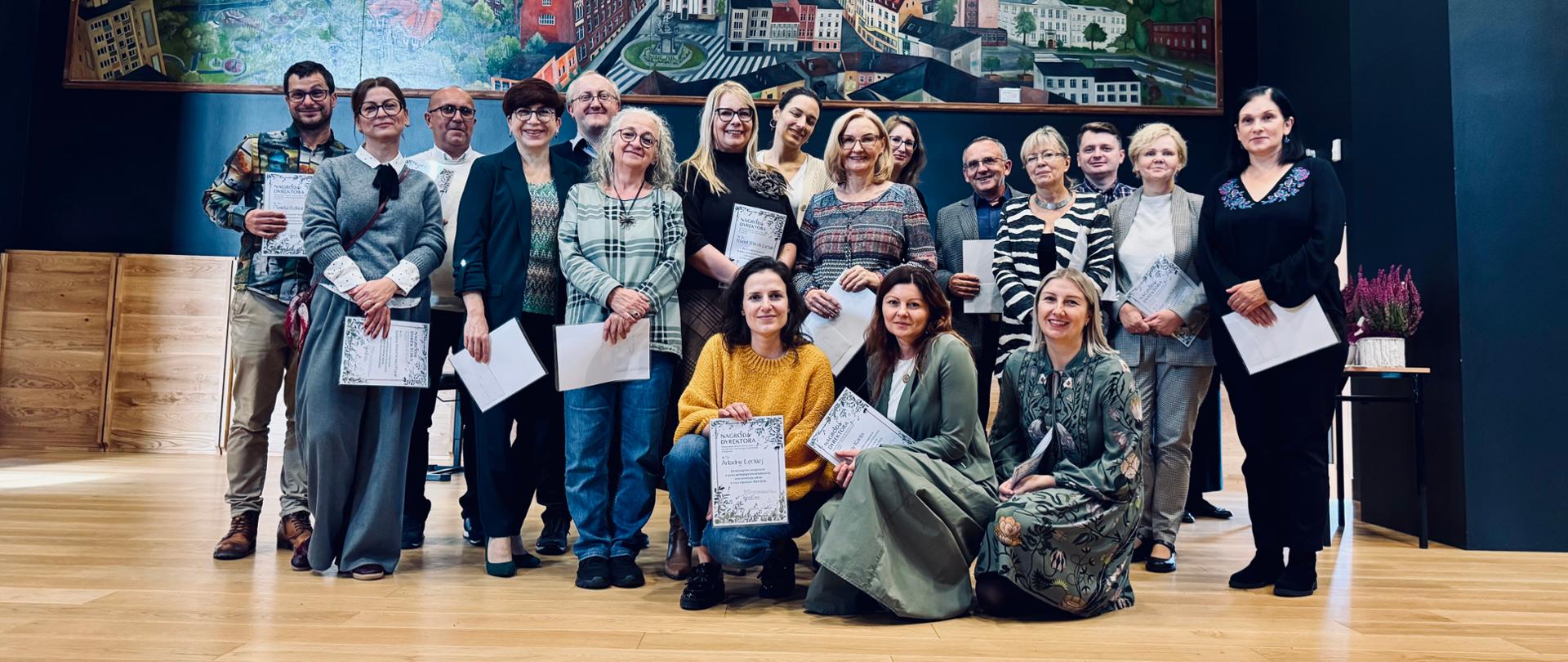 Grupa nauczycieli ustawiona w trzech rzędach na scenie sali koncertowej naszej szkoły. Pierwszy rząd - 3 osoby kucające (od lewej: Ariadna Łęcka, Joanna Kania i Anna Brachman), w drugim rzędzie - 8 osób stojących (od lewej: Justyna Dzierbicka-Paryl, Katarzyna Leśnik, Grażyna Hejno, Ksenia Więcek-Łuczak, Gizela Fritz, Joanna Maier, Katarzyna Balon i Aleksandra Skotarek. Trzeci rząd - 7 osób (od lewej: Paweł Szklarski, Grzegorz Grzegorzek, Artur Hes, Paulina Kulej, Barbara Bytner, Marceli Kubica i Krzysztof Maślanka. Wszyscy nauczyciele ubrani w kolorowe stroje, uśmiechnięci i trzymający w dłoniach dyplomy.