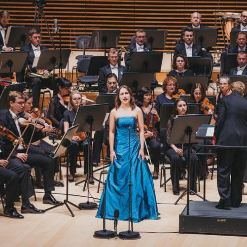 Kobieta śpiewa na estradzie, stojąc przed orkiestrą.