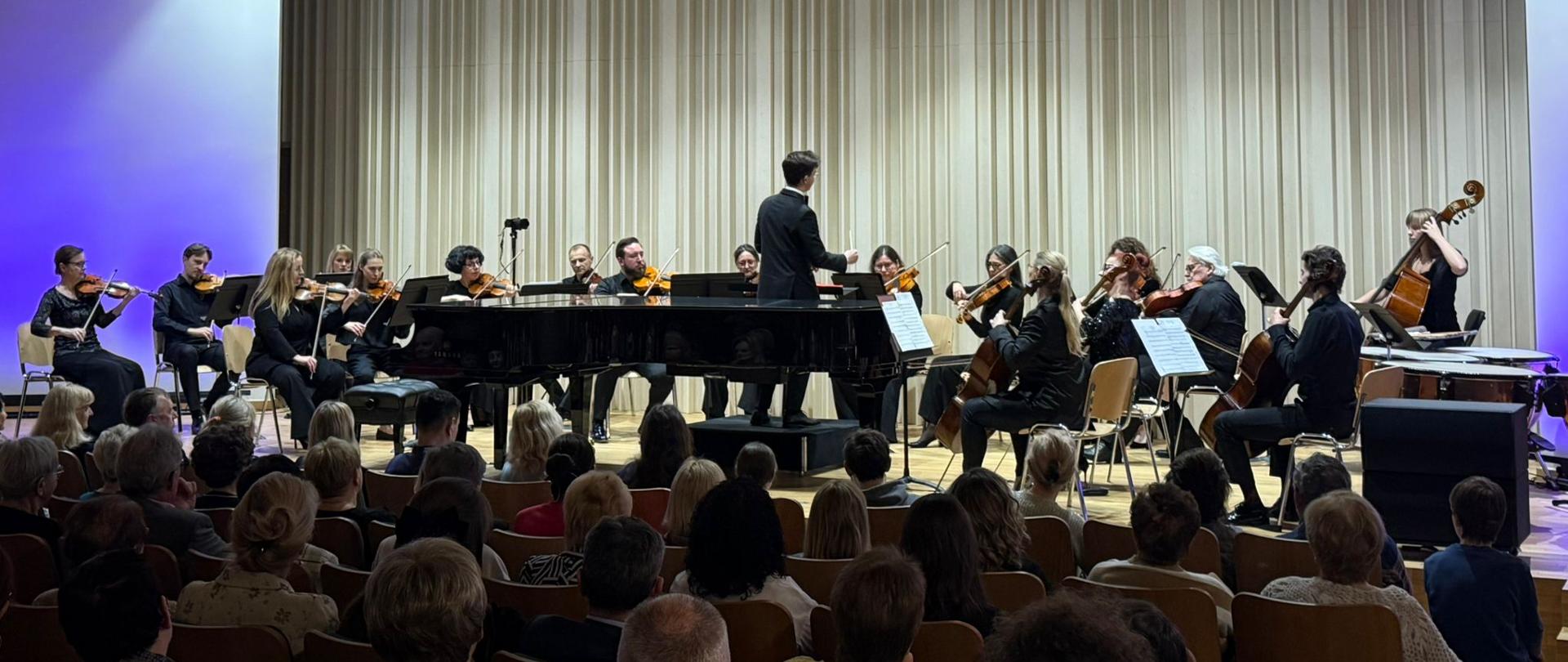 Na scenie licznie duża grupa muzyków, którzy siedzą na krzesłach i grają na instrumentach (skrzypce, wiolonczele, kontrabas, kotły). Przed nimi mężczyzna- dyrygent ubrany na czarno. W centralnej części sceny czarny fortepian. U dołu zdjęcia licznie zgromadzona publiczność. Zdjęcie zostało wykonane podczas koncertu Polskiej Filharmonii Kameralnej Sopot. Tło obrazka w odcieniach beżu z niebieskimi akcentami.