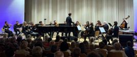 Na scenie licznie duża grupa muzyków, którzy siedzą na krzesłach i grają na instrumentach (skrzypce, wiolonczele, kontrabas, kotły). Przed nimi mężczyzna- dyrygent ubrany na czarno. W centralnej części sceny czarny fortepian. U dołu zdjęcia licznie zgromadzona publiczność. Zdjęcie zostało wykonane podczas koncertu Polskiej Filharmonii Kameralnej Sopot. Tło obrazka w odcieniach beżu z niebieskimi akcentami.