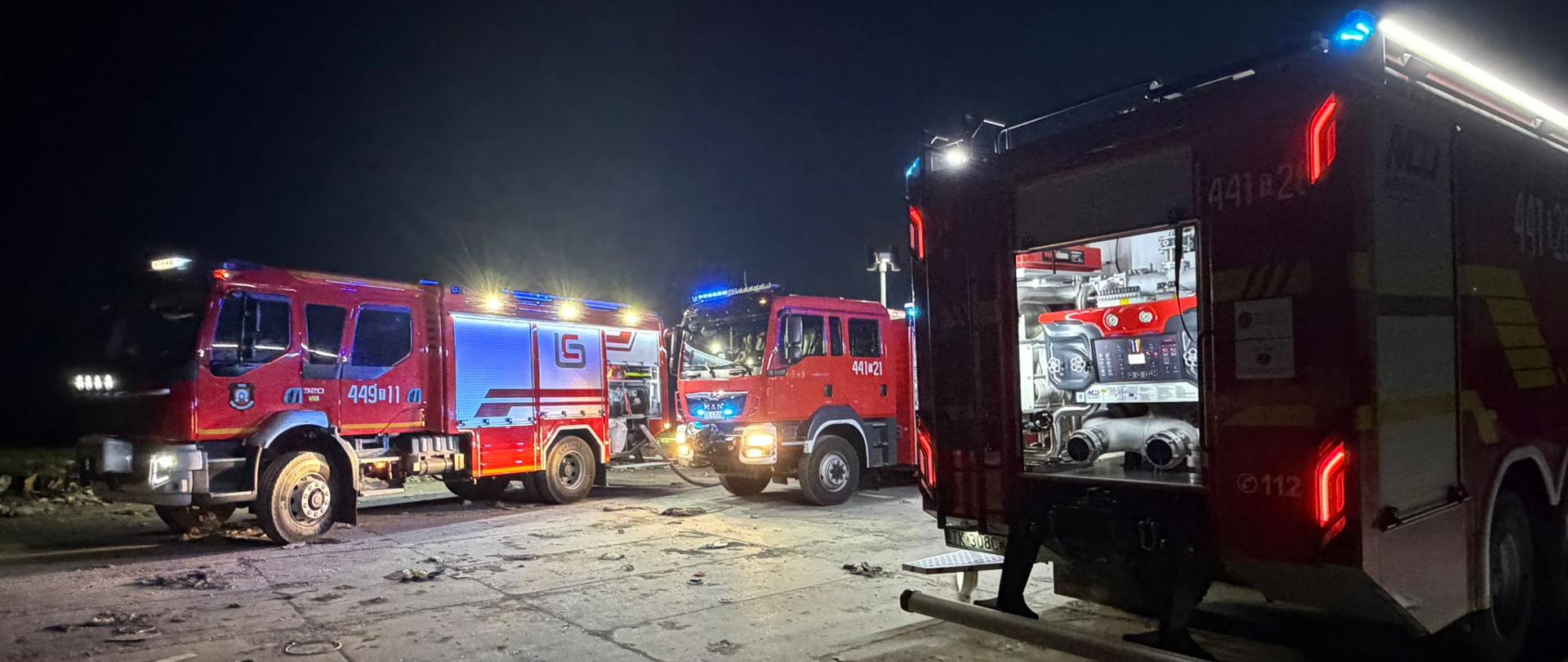 Na zdjęciu widać nocną scenę z udziałem kilku czerwonych wozów strażackich stojących na zniszczonej, zabrudzonej nawierzchni. Pojazdy mają włączone niebieskie i białe światła sygnalizacyjne, które oświetlają okolicę. Jeden z wozów ma otwartą tylną skrytkę, w której widoczny jest sprzęt strażacki. Całość sugeruje akcję ratunkową lub zabezpieczanie miejsca zdarzenia po zmroku.