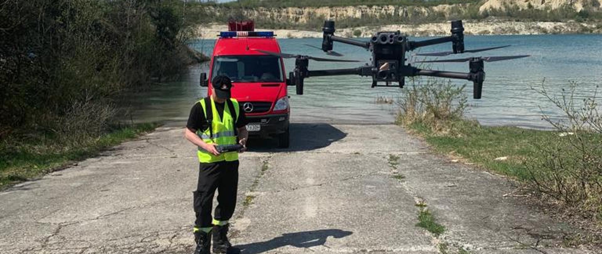 Zdjęcie wykonane w miejscowości Wierzbica na terenie zalanej kopalni żwiru. Na zdjęciu widać strażaka. Strażak w rękach trzyma sterowanie do drona. Przed nim w powietrzu unosi się dorn. Obok strażaka znajduje się również specjalna walizka transportowa umożliwiająca transport drona wraz z osprzętem. Za strażakiem w oddali widać pojazd pożarniczy w kolorze czerwonym. Pojazd typu bus. Służy do przewozu specjalistycznej grupy ratownictwa wodno - nurkowego. Strażak oraz pojazd znajdują się na utwardzonym zjeździe prowadzącym w stronę wody. Za nimi w oddali wiać zbiornik wody powstały z zalanej kopalni żwiru. 