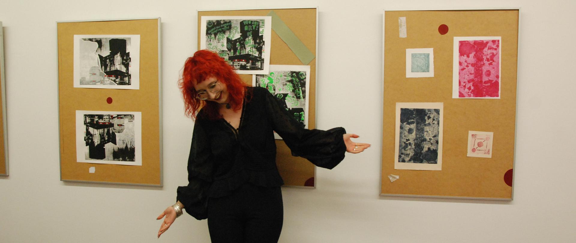 na białej ścianie wiszą tablice z grafikami, przed nimi stoi kłaniająca się kobieta