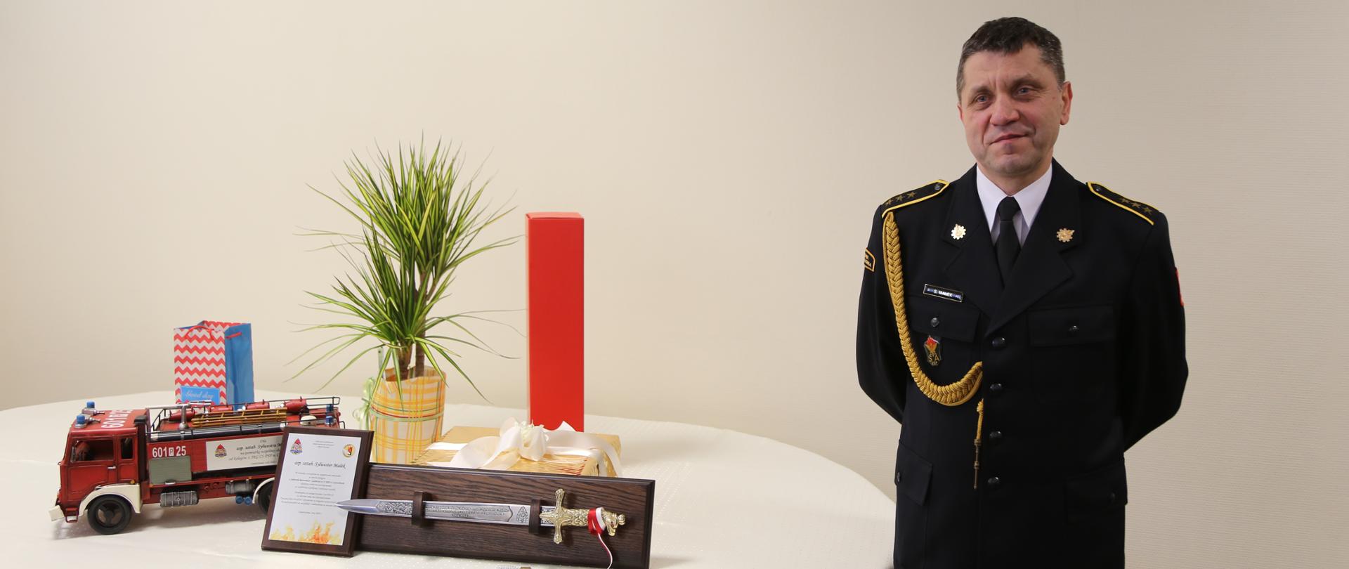 Na zdjęciu Pan asp. szt., z boku na stoliku okolicznościowe prezenty, które otrzymał tj.: kordzik umieszczony na ozdobnej desce, ryngraf oraz replika samochodu strażackiego Jelcz