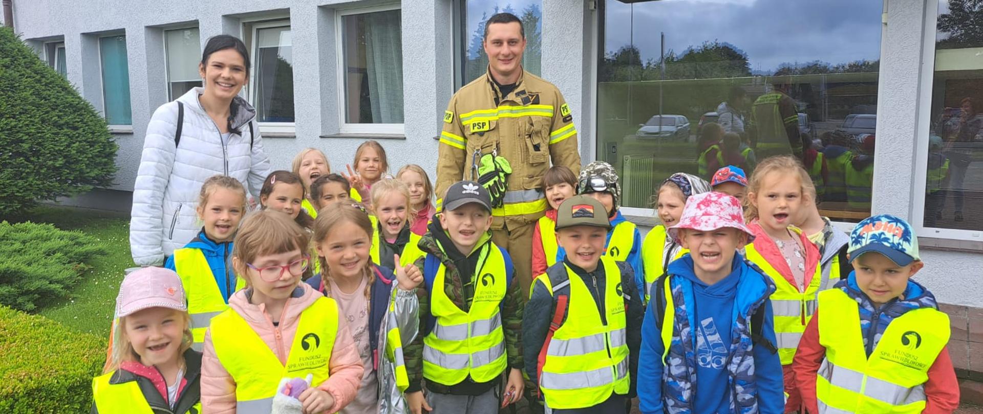 Zdjęcie przedstawia przedszkolaków ze strażakiem i opiekunem. Zdjęcie zrobione przed budynkiem komendy.