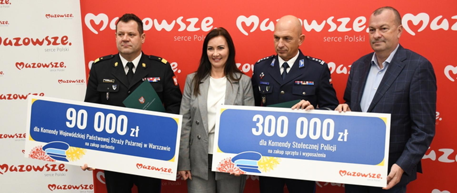 Na zdjęciu Mazowiecki Komendant Wojewódzki PSP - nadbryg. Artur Gonera, członkini Zarządu Województwa Mazowieckiego - Anna Brzezińska, Marszałek Województwa Mazowieckiego - Pan Adam Struzik i Zastępca Komendanta Stołecznego Policji - insp. Robert Frąckowiak. Stoją przed czerwonym banerem z białym napisem Mazowsze. Mazowiecki Komendant Wojewódzki PSP trzyma w ręku duży granatowy baner z białym napisem 90 000 zł, Zastępca Komendanta Stołecznego Policji baner z napisem 300 000 zł. 