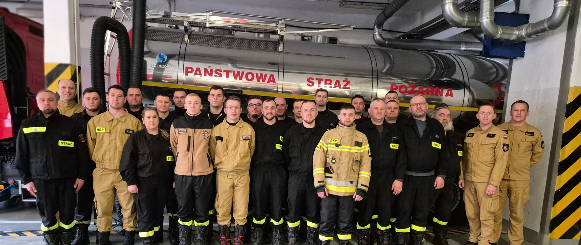 Strażacy stoją w szyku na garażu , w tle cysterna samochodowa z napisem Państwowa Straż Pożarna