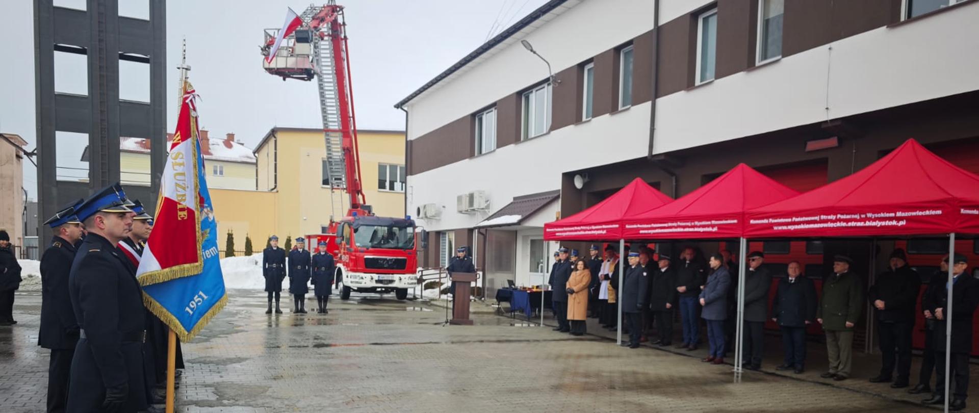 Na placu wewnętrznym Komendy Powiatowej PSP w Wysokiem Mazowieckiem odbyło się uroczyste włączenie do użytku pojazdów ratowniczo-gaśniczych oraz odprawa roczna podsumowująca działalność jednostek ochrony przeciwpożarowej powiatu wysokomazowieckiego za 2025 rok - plac wewnętrzny, zaproszeni goście, poczet sztandarowy i komendant powiatowy PSP przy mównicy. W tle wspinalnia.