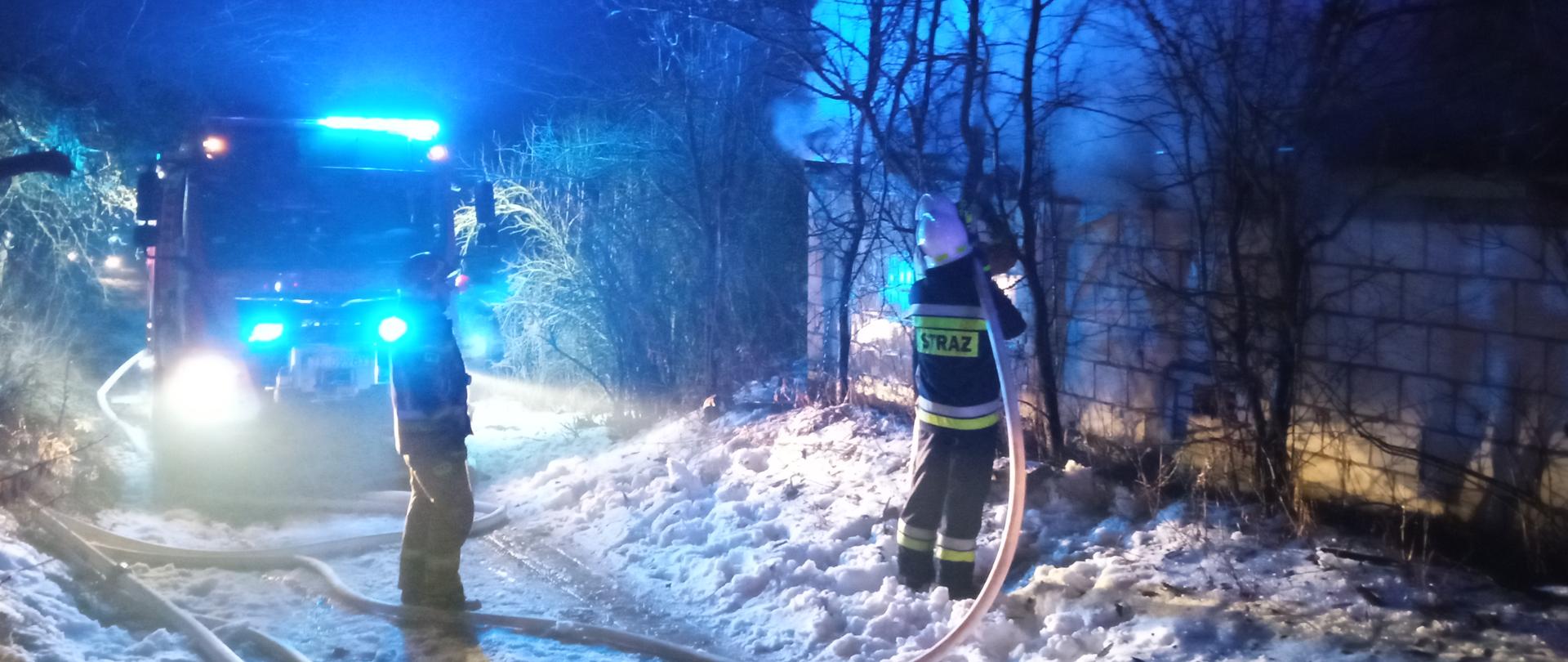 Zdjęcie zrobione w nocy. Na pierwszym planie dwóch strażaków w ubraniach specjalnych. Jeden trzyma węża i gasi pożar. Drugi asekuruje go. W tle stoi samochód strażacki na sygnałach błyskowych. Z budynku po prawej wydobywa się dym.