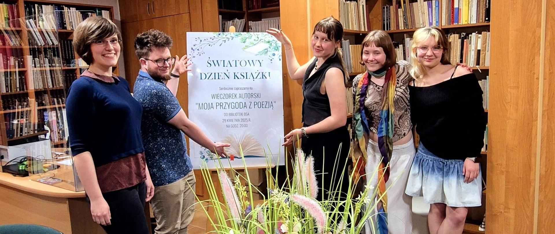 Zdjęcie wykonane w bibliotece. Pięcioro młodych roześmianych ludzi pozuje z plakatem ŚWIATOWY DZIEŃ KSIĄŻKI, za nimi są regały z książkami, przed nimi stół nakryty zielonym obrusem, a nim są ozdoby roślinne, serwetki, szklanki