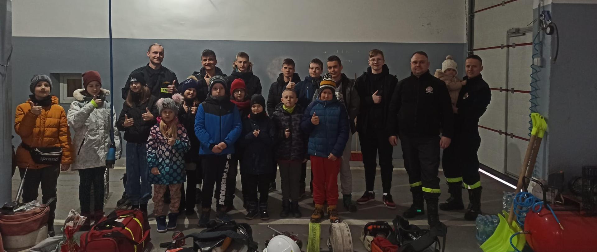 W garażu bojowym remizy strażackiej, biało szare ściany, stoi grupa dzieci ze strażakami. Na dole zdjęcia sprzęt strażacki.