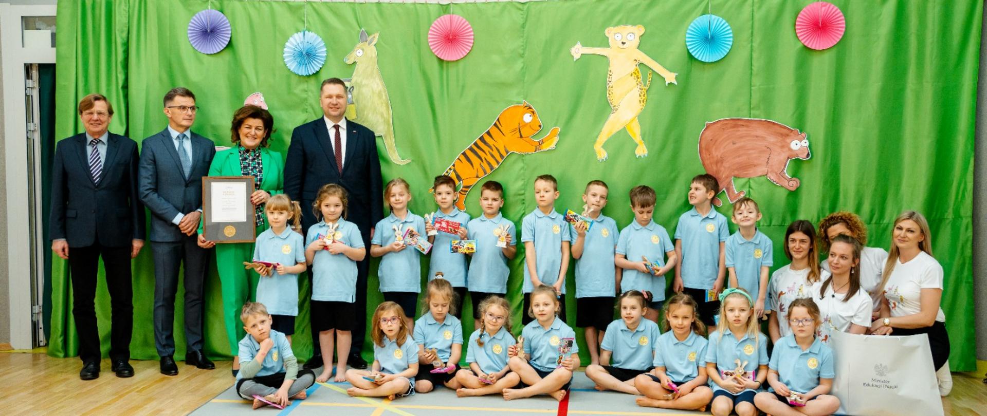 Na sali pod zieloną ścianą siedzi grupa jasnoniebiesko ubranych dzieci, za nimi stoi kilkoro dorosłych.