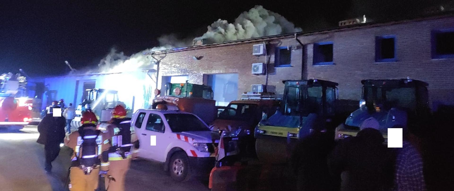 Zdjęcie zrobione na tle budynku magazynowego. Przed budynkiem stoi samochód osobowy typu pickup z zamontowanym pługiem, bus oraz dwa walce drogowe. Przed samochodem osobowym dwóch strażaków w umundurowaniu specjalnym, w hełmach oraz z butlami powietrznymi na plecach. Z lewej strony tył samochodu pożarniczego typu drabina. Z budynku magazynowego, przez dach wydobywają się kłęby dymu.