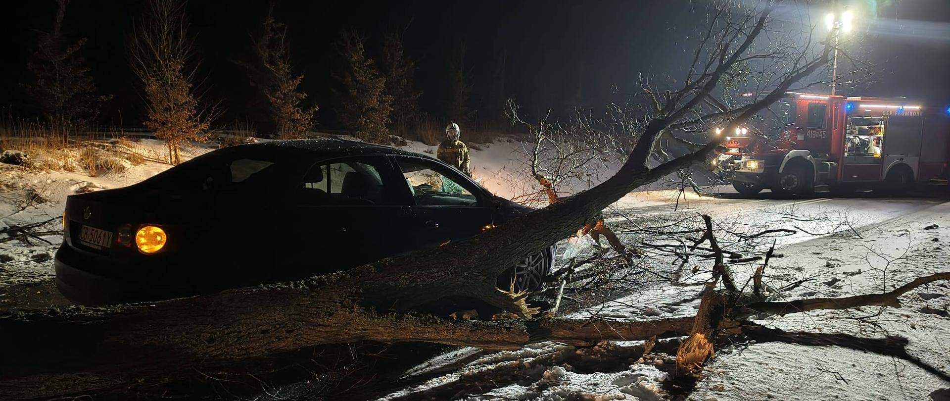 Zdjęcie zrobione nocą. Auto osobowe stoi na drodze przy złamanym drzewie, drzewo uderzyło w samochód. Z prawej strony u góry stoi samochód strażacki oświata teren zdarzenia.