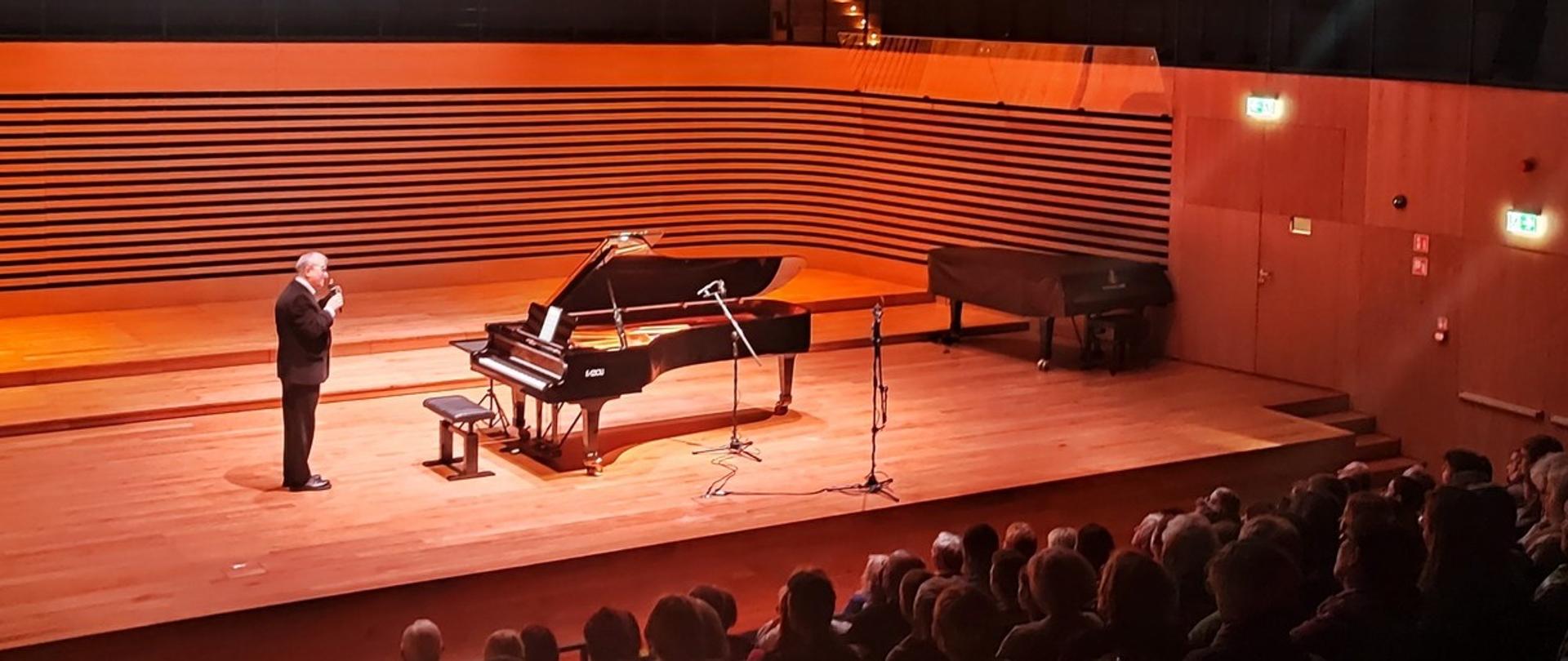 Artysta stoi przy fortepianie i mówi do mikrofonu, z przodu przed sceną widać publiczność