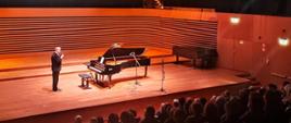 Artysta stoi przy fortepianie i mówi do mikrofonu, z przodu przed sceną widać publiczność