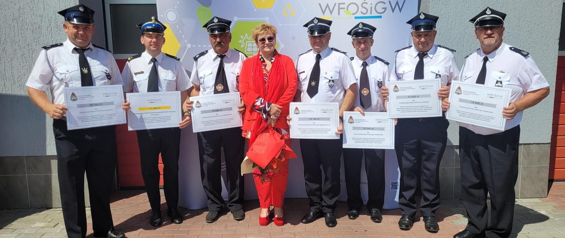 Wręczenie promes. Plac przed Komendą Miejską PSP w Koninie. Na tle baneru reklamowego WFOŚiGW w Poznaniu stoją strażacy z powiatu rawickiego, którzy otrzymali promesy. W środku pani prezes WFOŚiGW w czerwonym kostiumie. Strażacy występują w umundurowaniu letnim (biała koszula z krótkim rękawem). 