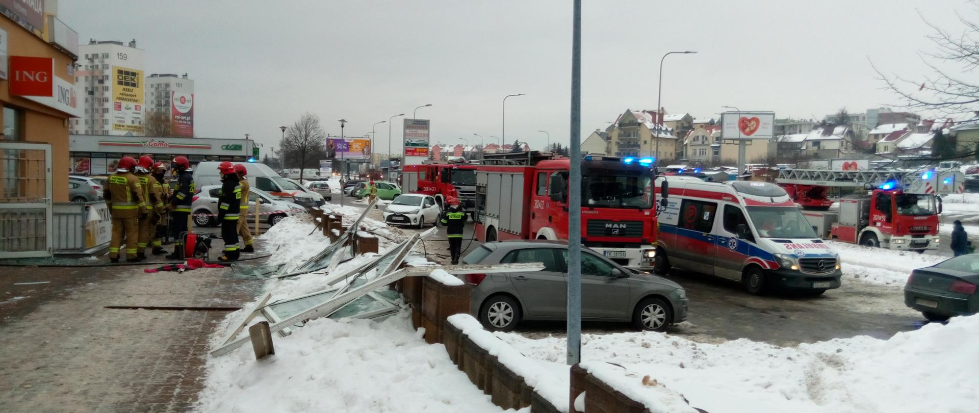 Na zdjęciu widać grupę strażaków stojącą przed budynkiem pralni. Na chodniku znajduje się wybite okno. Zaraz za chodnikiem widać samochód strażacki, karetkę pogotowia oraz pozostałe samochody osobowe znajdujące się na parkingu.