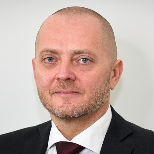 Mężczyzna w ciemnej marynarce, białej koszuli, bordowym krawacie, z krótkim zarostem.