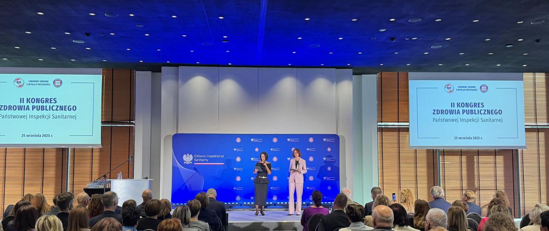 Zdjęcie przedstawia scenę konferencyjną podczas II Kongresu Zdrowia Publicznego Państwowej Inspekcji Sanitarnej, który odbył się 25 września 2025 roku. Wydarzenie odbywa się w przestronnej sali konferencyjnej, przed liczną publicznością.
Na scenie stoją dwie prowadzące – jedna w czarnym stroju, druga w jasnoróżowym garniturze. Znajdują się przed niebieskim tłem z logotypami Głównego Inspektoratu Sanitarnego (GIS) oraz innych instytucji współorganizujących kongres. Nad sceną umieszczono dwa ekrany, po jednym z każdej strony, na których widnieje:
II KONGRES ZDROWIA PUBLICZNEGO
Państwowej Inspekcji Sanitarnej
25 września 2025 r.
W tle widać duże okna z żaluzjami, które przepuszczają naturalne światło. Na pierwszym planie siedzi publiczność – uczestnicy kongresu. Atmosfera jest oficjalna i profesjonalna, co sugeruje rangę i znaczenie wydarzenia dla sektora zdrowia publicznego w Polsce.