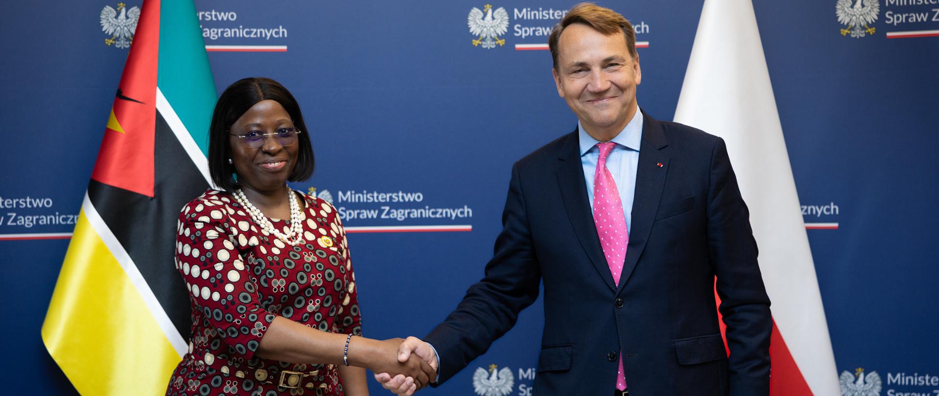 Maria Manuela dos Santos Lucas i Radosław Sikorski podają sobie ręce na tle granatowej ścianki z logotypami Ministerstwa Spraw Zagranicznych RP oraz flag Mozambiku i Polski. Oboje uśmiechają się do zdjęcia. Fotografia dokumentuje oficjalne spotkanie.