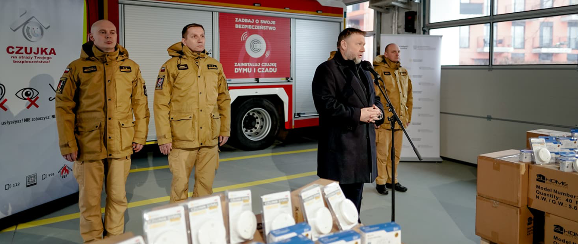 Minister Marcin Kierwiński i przedstawiciele PSP podczas konferencji prasowej. W tle samochód strażacki, a po bokach wyeksponowane na kartonach czujki dymu i tlenku węgla.