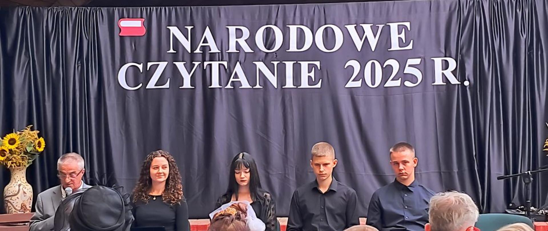 Dyrektor szkoły wraz z czwórką uczniów siedzą na tle kurtyny z napisem "Narodowe czytanie 2025R"