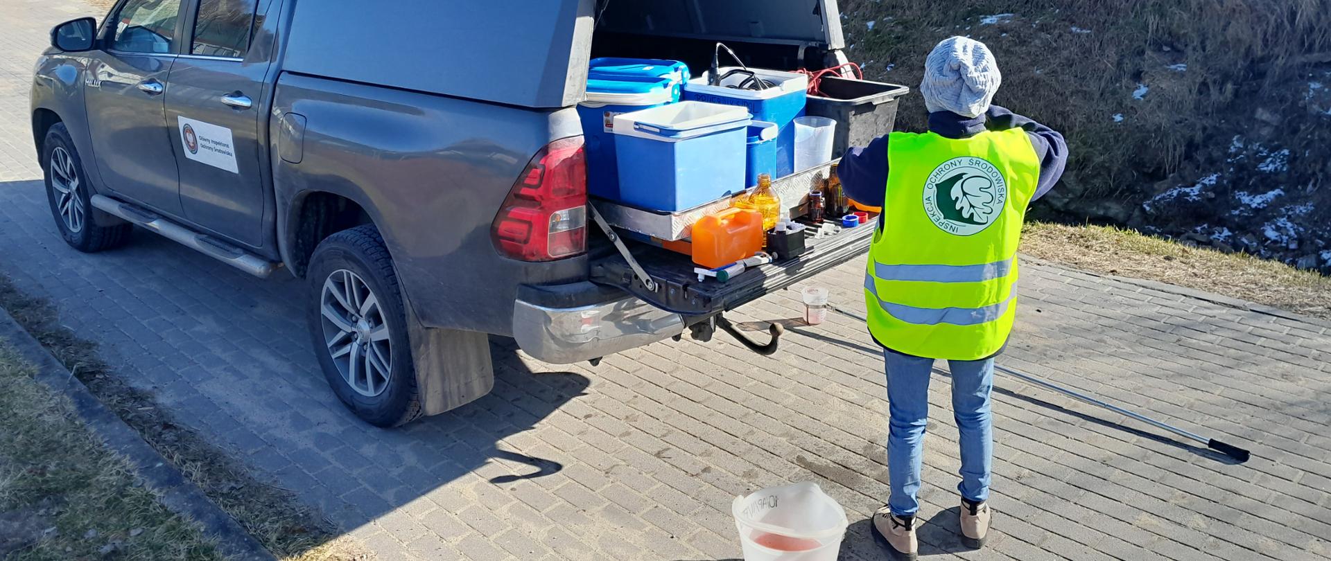Szary samochód terenowy z sprzętem laboratoryjnym. Wiadro z czerwoną wodą z rowu. W tle łąki i las.