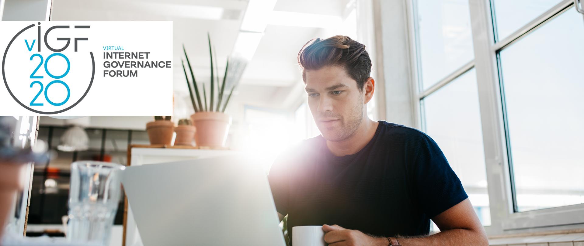 Młody mężczyzna przed białym laptopem, w dłoni trzyma biały kubek. W tle wnętrze pomieszczenia, widoczne rośliny doniczkowe, stojąca na stole szklanka, z tyły pada światło słoneczne, z prawej strony - okno.