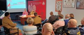 Zdjęcie przedstawia scenę ze spotkania lub narady w sali konferencyjnej z udziałem służb mundurowych i lokalnych władz. W centralnym punkcie, za biurkiem, siedzą dwie osoby: Prezydent w łososiowym swetrze oraz strażak w piaskowym mundurze Państwowej Straży Pożarnej (PSP). Po lewej stronie, na dużym ekranie, wyświetlana jest prezentacja z logo KSRG (Krajowy System Ratowniczo-Gaśniczy) i napisem "STRAŻ POŻARNA".
Przed biurkiem, skierowana tyłem do osoby fotografującej, siedzi grupa słuchaczy. Wśród nich widać osoby cywilne oraz funkcjonariuszy w mundurach, w tym strażaków PSP oraz, na pierwszym planie, osobę z napisem "STRAŻ MIEJSKA". Ściana za biurkiem jest ozdobiona dużym, czerwonym panelem z godłem Polski i logo PSP, a na prawej ścianie wiszą oprawione w ramki wykresy organizacyjne lub tablice pamiątkowe. Ujęcie to skupia się na interakcji i odbiorze informacji przez publiczność podczas oficjalnego spotkania.
