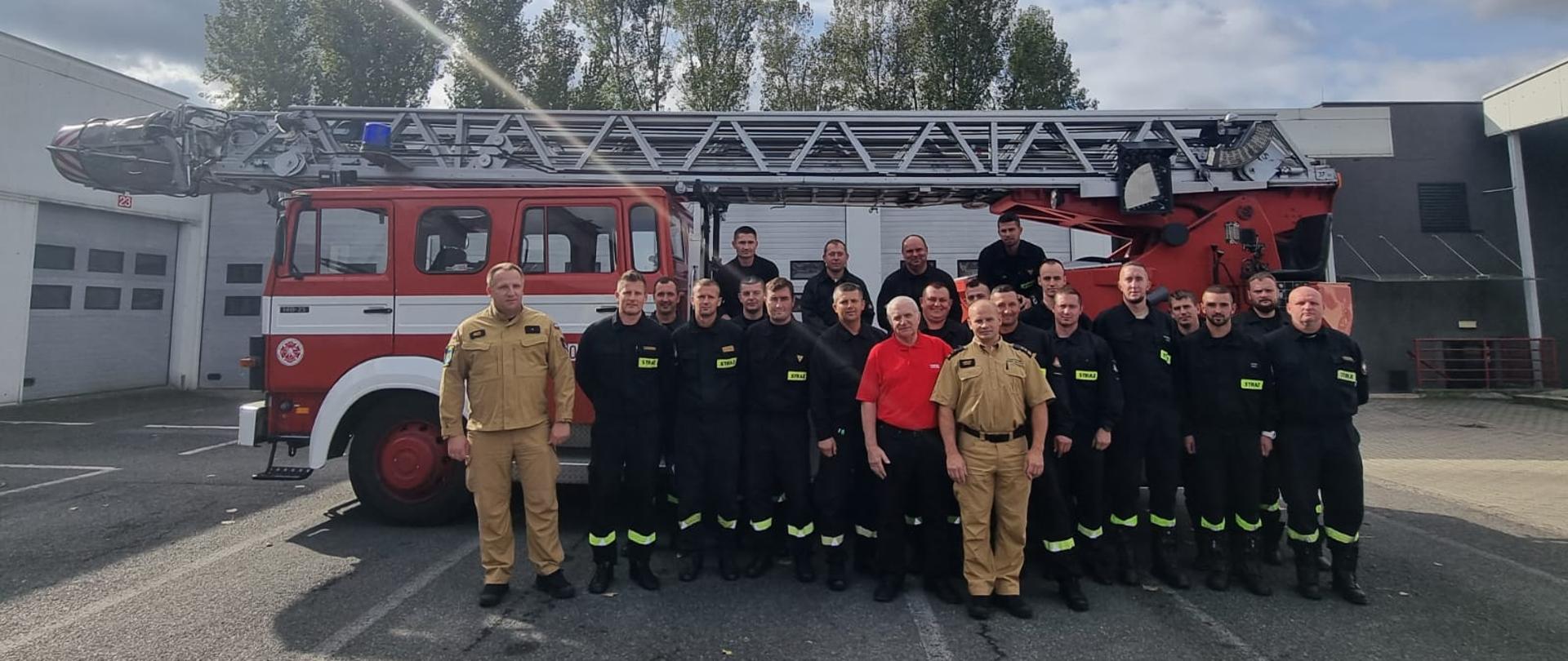 Wspólne zdjęcie grupowe uczestników szkolenia oraz wykładowców szkolenia specjalistycznego kierowców - operatorów samochodów z drabiną mechaniczną, które odbyły się w Ośrodku Szkolenia Komendy Wojewódzkiej PSP w Opolu. Strażacy stoją na tle samochodu specjalistycznego - drabiny mechanicznej na placu Komendy Wojewódzkiej PSP w Opolu.