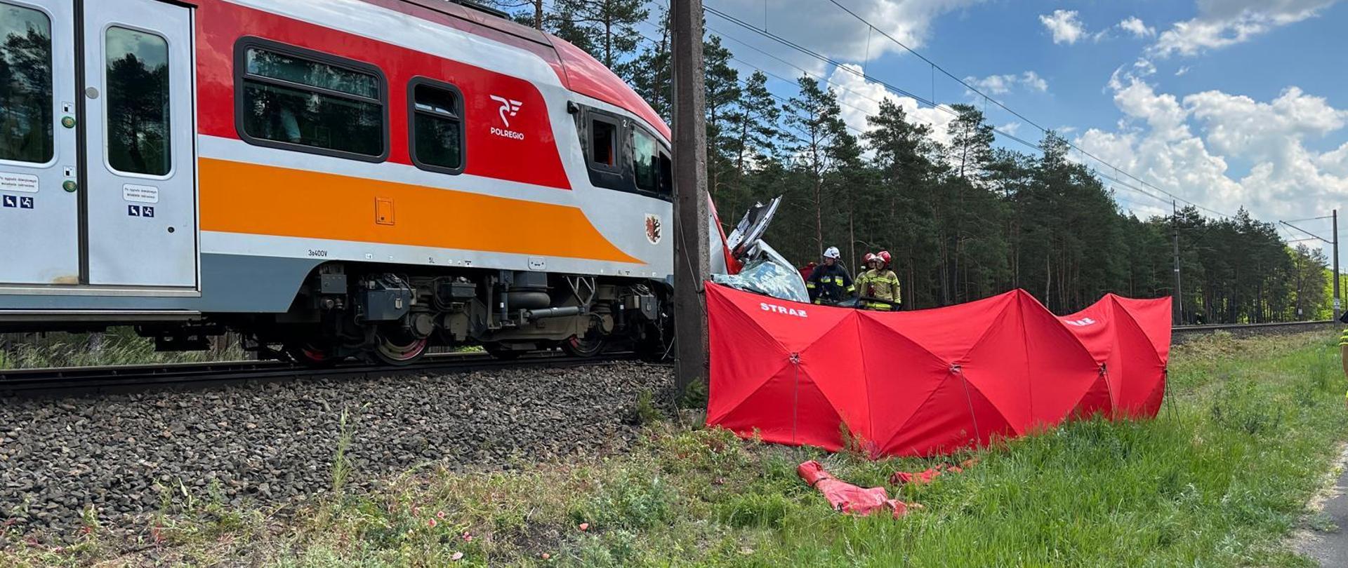 Pociąg a przed nim zniszczony samochód, obok stoi parawan z napisem straż.