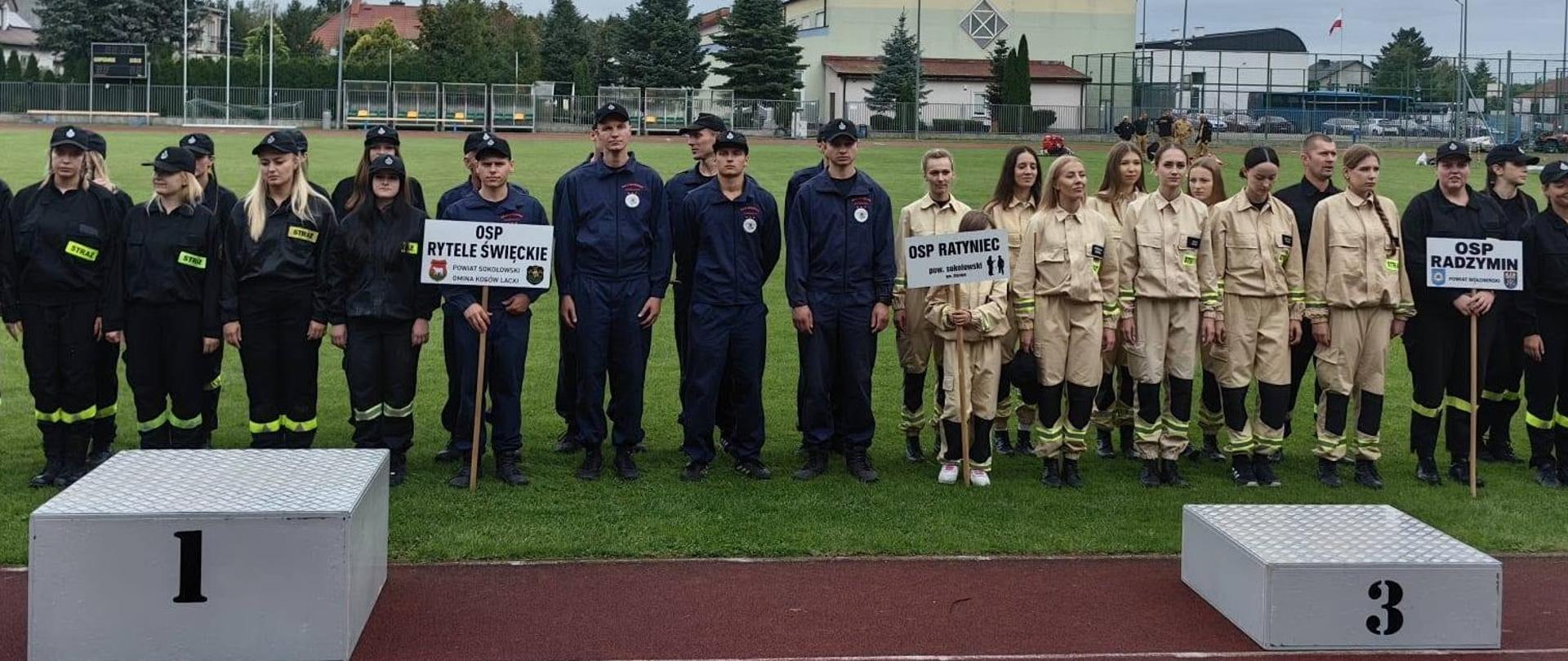 Szerokie ujęcie przedstawiające oficjalną zbiórkę drużyn na terenie boiska sportowego / bieżni lekkoatletycznej. Ustawienie: Na zdjęciu widoczne są w kolejności od lewej strony m.in. drużyny OSP Rytele Święckie (w granatowych dresach), OSP Ratyniec Stary (w piaskowych mundurach) oraz OSP Radzymin. Z przodu widoczne są podest podium z numerami 1 oraz 3. W tle rozciąga się murawa boiska oraz zabudowania w tle