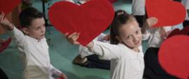 Na sali tańczą ubrane na biało dzieci, trzymają w rękach duże czerwone plakietki w kształcie serca.
