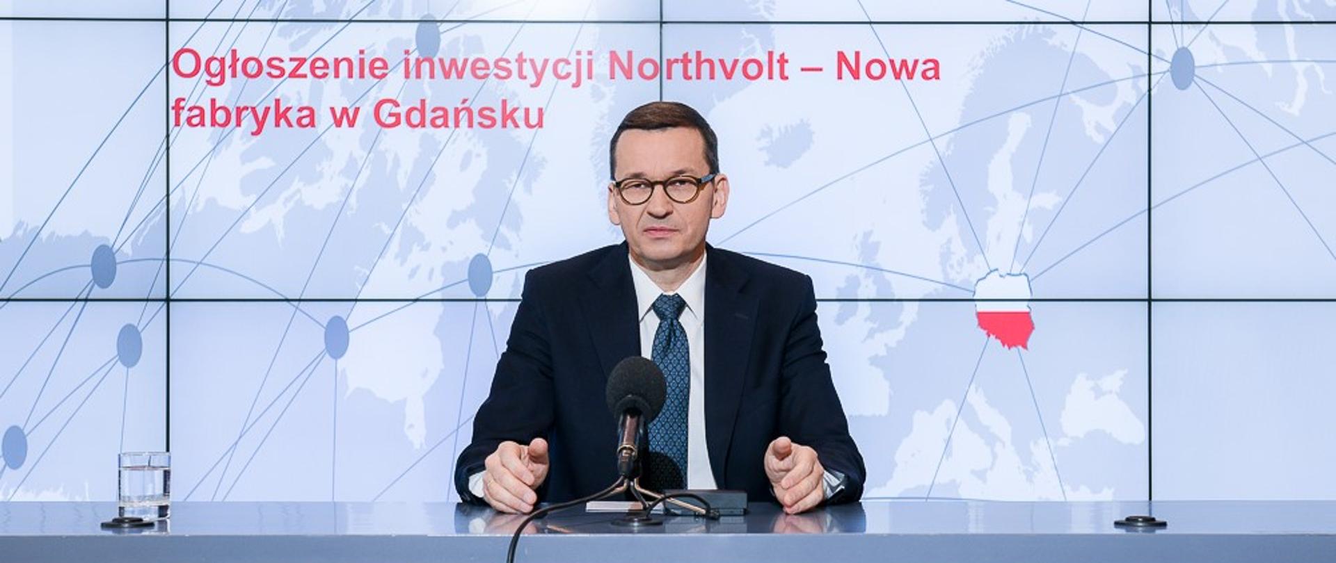 Premier Mateusz Morawiecki siedzi przy stole, na którym jest ustawiony mikrofon. Przed nim godło Polski. W tle ścianka multimedialna z napisem "Ogłoszenie inwestycji Northvolt - Nowa fabryka w Gdańsku".