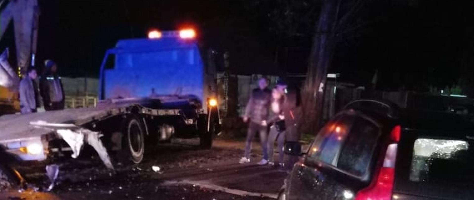 Na zdjęciu samochód osobowy w kolorze czarnym po uderzeniu w autolawetę. Kabina lawety w kolorze niebieskim. Pomiędzy pojazdami stoją dwie osoby na jezdni. Zdjęcie wykonane porą nocną.