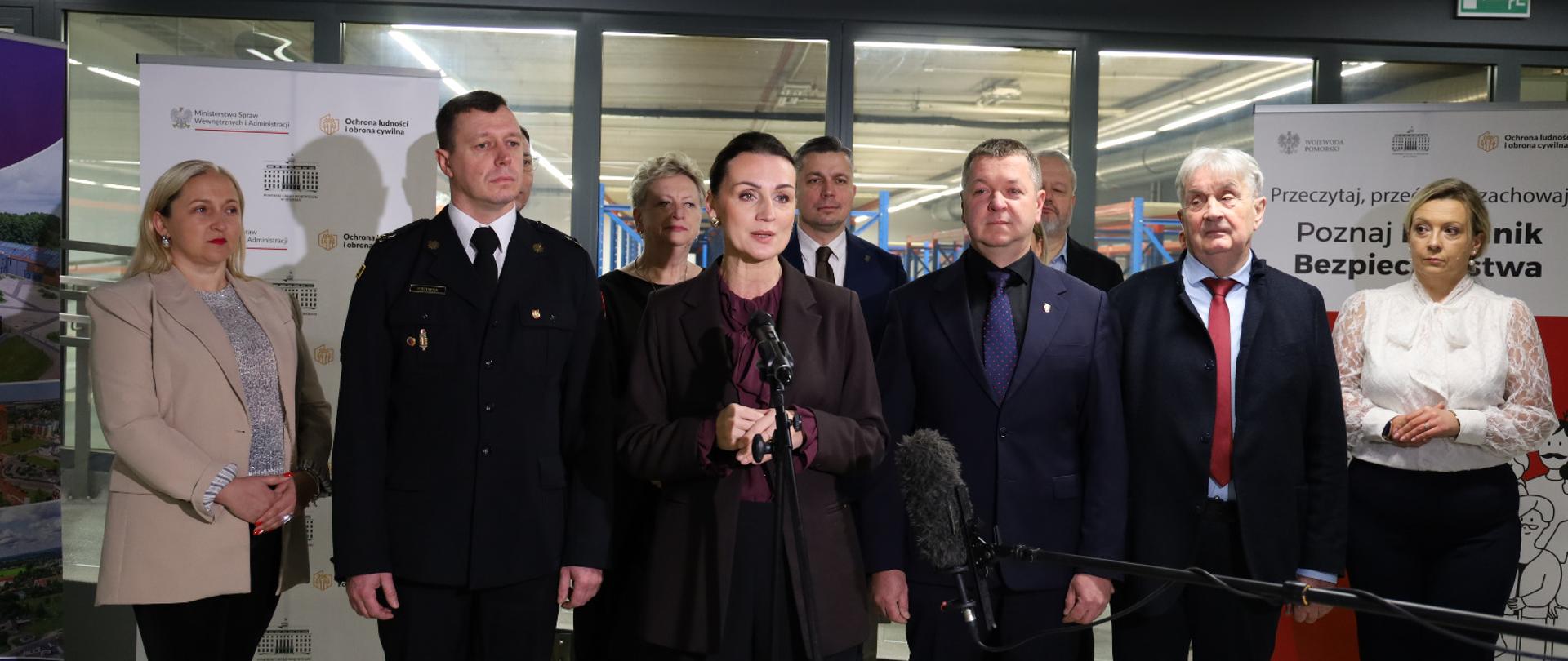 Grupa ludzi stojąca w magazynie. Przy mikrofonie stoi kobieta (wojewoda).