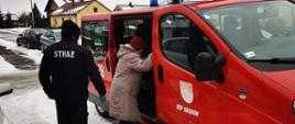 Do małego samochodu strażackiego wsiada starsza kobieta. Za kobietą stoi strażak, starający się jej pomóc wejść do pojazdu. Na chodniku leży śnieg. W oddali widoczne są zabudowania jednorodzinne oraz pojazdy ustawione przy ulicy. 
