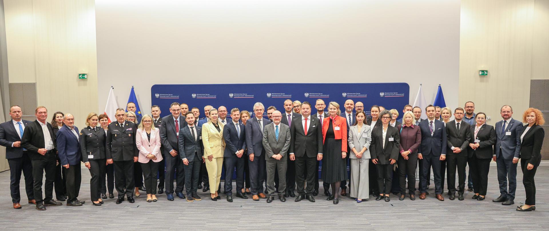 Grupa kilkudziesięciu osób, w tym przedstawiciele służb mundurowych i urzędnicy w garniturach, stoi wspólnie do zdjęcia grupowego na tle ściany z logo Ministerstwa Spraw Wewnętrznych i Administracji. W tle widoczne są flagi Polski i Unii Europejskiej. Fotografia wykonana podczas spotkania lub konferencji międzynarodowej.