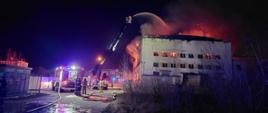 W nocy, budynek hali przemysłowej objęty pożarem. Pożar gaszą strażacy z kosza wysuniętej drabiny samochodowej. Przed budynkiem hali stoi również inny samochód ratowniczo-gaśniczy, obok którego stoją strażacy. 
