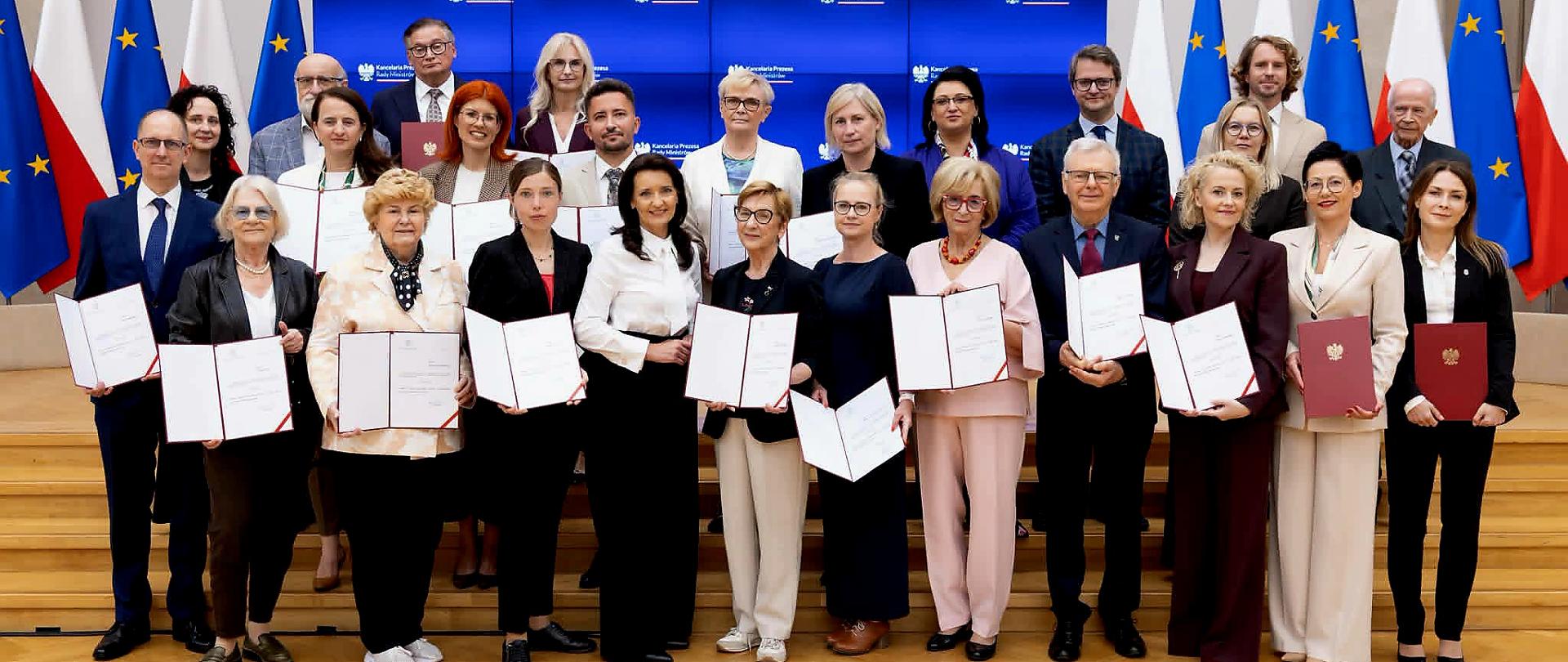 Zdjęcie grupowe około 25 osób pozujących w sali konferencyjnej przed ekranami z logo Kancelarii Prezesa Rady Ministrów. W tle stoją naprzemiennie flagi Polski i Unii Europejskiej. Część osób trzyma otwarte teczki z dokumentami lub czerwone teczki z godłem Polski.